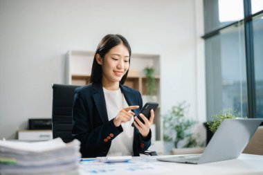 Genç Asyalı kadın, gülümsüyor ve ofis ortamında masasında çalışıyor. Bir iş elbisesi giyiyor ve dizüstü bilgisayar kullanırken elinde belgeler tutuyor.