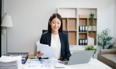 Genç Asyalı kadın, gülümsüyor ve ofis ortamında masasında çalışıyor. Bir iş elbisesi giyiyor ve dizüstü bilgisayar kullanırken elinde belgeler tutuyor..