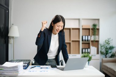 Genç Asyalı kadın, gülümsüyor ve ofis ortamında masasında çalışıyor. Bir iş elbisesi giyiyor ve dizüstü bilgisayar kullanırken elinde belgeler tutuyor..