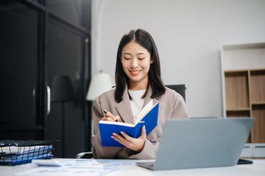 Genç Asyalı kadın, gülümsüyor ve ofis ortamında masasında çalışıyor. Bir iş elbisesi giyiyor ve dizüstü bilgisayar kullanırken elinde belgeler tutuyor..