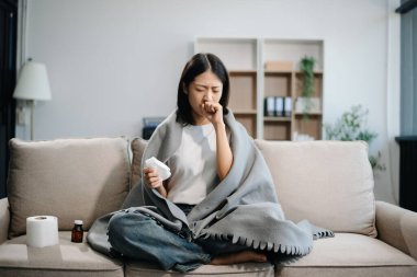 Kanepede battaniyeyle kaplanmış, grip belirtileri gösteren genç bir Asyalı kadın. Hastalık, sağlık ve iyileşme kavramı. Evde kal, sağlıklı kal.