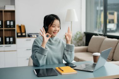 Çekici mutlu Asyalı öğrenci üniversite kütüphanesinde okuyor, masada oturuyor, bilgisayarını, tabletini ve kulaklığını kullanıyor ve görüntülü konuşma yapıyor..