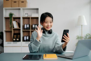 Çekici mutlu Asyalı öğrenci üniversite kütüphanesinde okuyor, masada oturuyor, bilgisayarını, tabletini ve kulaklığını kullanıyor ve görüntülü konuşma yapıyor..