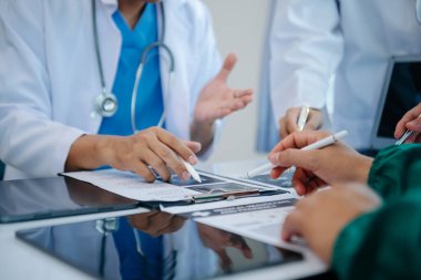 Tıbbi ekip, beyaz laboratuvar önlüklü ve ameliyat önlüklü doktorlarla toplantı yapıyor. Masada oturmuş, internette çalışan hastaları tartışıyorlar. Tıp endüstrisinde bilgisayar kullanıyorlar.