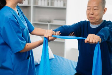 Asyalı fizyoterapist, yaşlı bir adamın egzersiz sırasında kollarını esnetmesine yardım ediyor. Hasta ile antrenman sırasında elinde dambıl vardı. Yatakta sırt sorunları vardı.