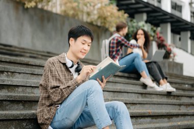 Üç genç üniversite öğrencisi, kampüs parkında arkadaşlarıyla çimde otururken rahatlarken kitap okuyor. Sabah eğitim kavramı