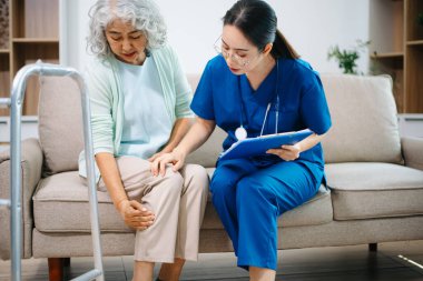 Asyalı hemşire evdeki koltukta oturan yaşlı bir kadının diz ve bacak ağrısını inceliyor. Eklem ağrısı, eklem iltihabı, teşhis, hareket kabiliyeti ve modern ev sağlığı hizmetlerine odaklanıyor..