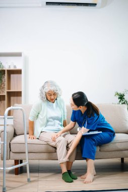 Asyalı hemşire evdeki koltukta oturan yaşlı bir kadının diz ve bacak ağrısını inceliyor. Eklem ağrısı, eklem iltihabı, teşhis, hareket kabiliyeti ve modern ev sağlığı hizmetlerine odaklanıyor..