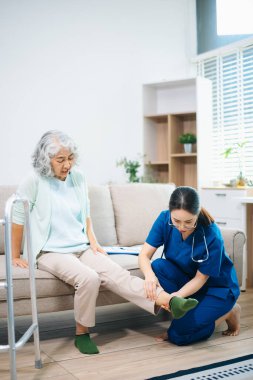 Asyalı hemşire evdeki koltukta oturan yaşlı bir kadının diz ve bacak ağrısını inceliyor. Eklem ağrısı, eklem iltihabı, teşhis, hareket kabiliyeti ve modern ev sağlığı hizmetlerine odaklanıyor..