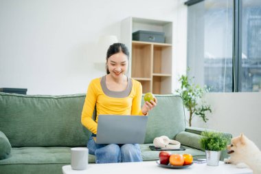 Modern evdeki kanepede otururken akıllı telefon ve tablet kullanan genç Asyalı kadın.