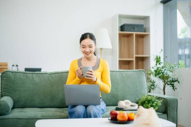 Modern evdeki kanepede otururken akıllı telefon ve tablet kullanan genç Asyalı kadın.