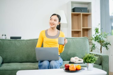 Modern evdeki kanepede otururken akıllı telefon ve tablet kullanan genç Asyalı kadın.