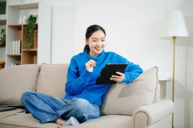 Genç Asyalı kadın akıllı telefon ve tablet kullanarak evdeki kanepede dinleniyor. Neşeli online alışveriş, dijital yaşam tarzı, ve rahat modern bir yaşam alanında kablosuz teknoloji..