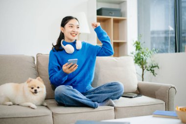 Kanepede oturan genç bir kadın evinde şirin beyaz bir köpek yavrusuyla dizüstü bilgisayar kullanıyor. Konforlu modern oturma odası