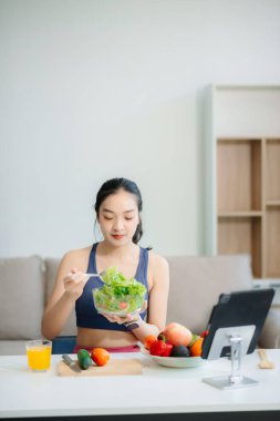 Neşeli bir kadın olarak sağlıklı beslenme, taze üretim ve sağlığın canlı bir sahnesi evde teknolojiyle uğraşırken salata ve meyvelerin tadını çıkarıyor.