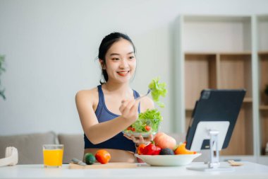 Neşeli bir kadın olarak sağlıklı beslenme, taze üretim ve sağlığın canlı bir sahnesi evde teknolojiyle uğraşırken salata ve meyvelerin tadını çıkarıyor.