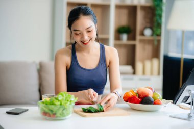 Neşeli bir kadın olarak sağlıklı beslenme, taze üretim ve sağlığın canlı bir sahnesi evde teknolojiyle uğraşırken salata ve meyvelerin tadını çıkarıyor.