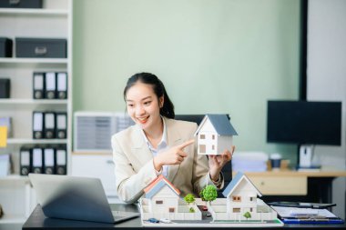 Modern ofis danışmanlığında Asyalı bir emlakçı müşterilere danışmanlık yapıyor. Dizüstü bilgisayarlı, tabletli ve ev modelli mülkler sergiliyor. Profesyonel, güvenilir.