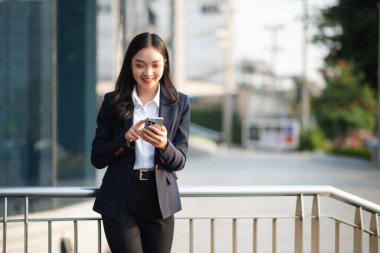 Ofisin dışında elinde not defteri ve tabletle gülümseyen kendine güvenen Asyalı kadın. İş merkezi arka planında büyük şehir.