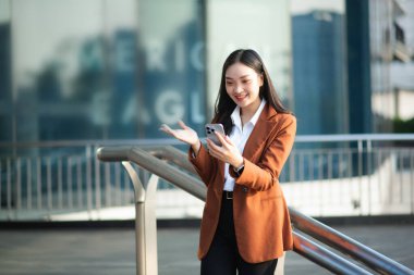 Ofisin dışında elinde not defteri ve tabletle gülümseyen kendine güvenen Asyalı kadın. İş merkezi arka planında büyük şehir.