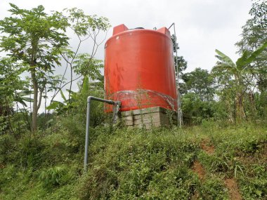 Su depolama ve sulama için kullanılan yeşil kırsal alanda taşan büyük bir kırmızı su tankı. Su arzı ve tarım altyapısını temsil ediyor.
