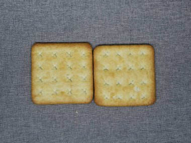 Koyu gri tekstil arka planında blok şeklinde dizilmiş iki kare tuzlu kraker bisküvisinin yukarıdan aşağı görüntüsü. Bu çıtır çıtır, altın aperatifler yemek ve kahvaltı kavramları için mükemmeldir..