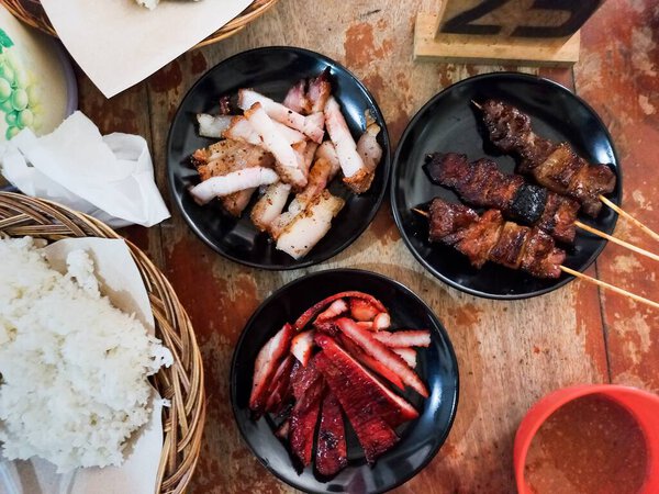 Authentic Asian pork feast: Samcan or crispy pork belly, bright red Char Siu or BBQ pork, and savory pork satay skewers served with steamed rice on a wooden table.