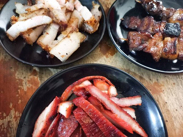 Authentic Asian pork feast: Samcan or crispy pork belly, bright red Char Siu or BBQ pork, and savory pork satay skewers served with steamed rice on a wooden table.