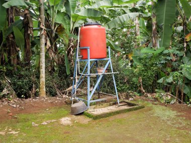 Muz ağaçlarıyla çevrili, kırsal ve tropik bir çevrede su depolamak için kullanılan mavi çelik bir tezgahta yükseltilmiş turuncu plastik bir su tankı..