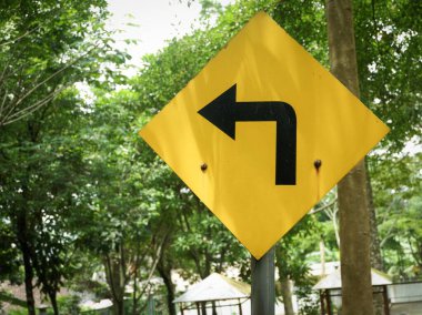 Elmas şekilli, sarı trafik uyarısı. İleride keskin ya da zorunlu bir dönüş olduğunu gösteriyor. Metal bir direğe kurulmuş, yemyeşil bir orman ya da park arka planında..