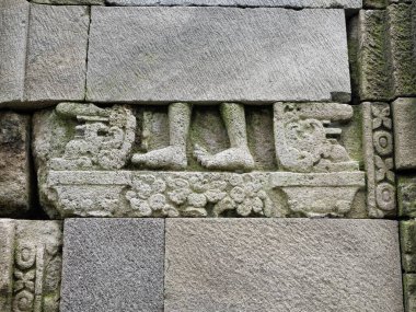 Yıpranmış gri volkanik taş duvarlar ayakların detaylı bir şekilde oyulduğunu ve doku çatlaklarında yosunların büyüdüğünü gösteriyor..