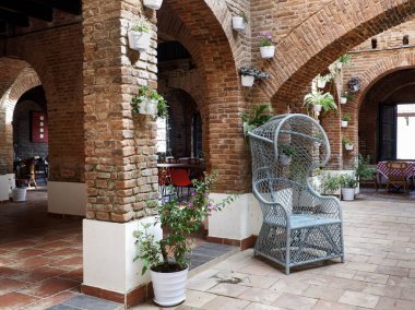 Açığa çıkmış tuğla kemer ve kolonlara sahip eski bir binanın iç manzarası, rustik örülmüş bir rattan sandalyeye ve saksı bitkilerine sahip. Sıcak, antika kafe tarzı..