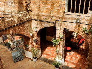 Terracotta tuğla kemerleri ve döşeme kiremitleriyle güneş ışığıyla aydınlatılmış iç avlu. Kırsal Akdeniz mimarisini ön plana çıkaran bir rattan sandalye ve saksı bitkisi.