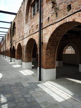 Yenilenmiş tuğla bina koridorunun kemer sütunlu uzun, perspektifli manzarası. Modern asfalt döşemeler, beyaz üsler ve güçlü gölgeler. Tarihi mimari.
