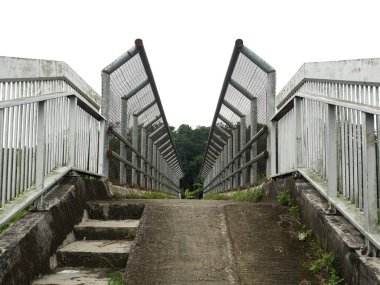 Merkezi beton bir rampa ve metal parmaklıklar ve tel örgülerle çerçevelenmiş yan basamakları olan yaya üstgeçit köprüsünün simetrik görüntüsü.