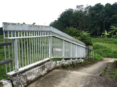 Beton bir yaya rampası boyunca uzanan endüstriyel metal köprü, verimli bir tropikal orman manzarasına dönüşüyor. Kırsal bir ortamda eskimiş mimari detaylar.