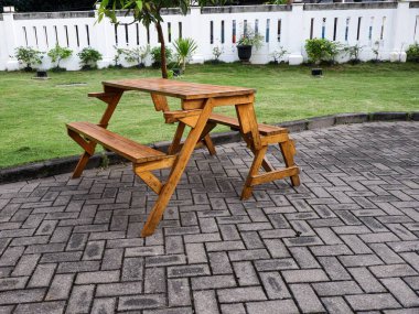 Ahşap katlanabilir bir piknik masası ve tuğladan bir verandadaki bank setinin yan açılı görüntüsü. Arka planda yeşil bir çimen, beyaz bahçe duvarı ve yemyeşil tropik ağaçlar yer alıyor..