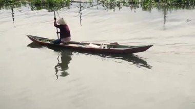 Geleneksel bir balıkçı günbatımında sakin bir bataklıkta tahta bir kayıkla kürek çeker. Sümbül ağaçları ve kırsal bir stistik ev sakin bir sulak arazinin altın suyuna yansıyor..