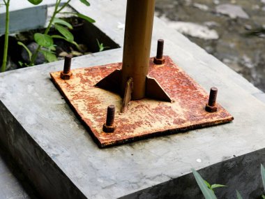 Beton bir temele sabitlenmiş paslı bir metal taban levhanın yakın çekimi. Korozyon, yıpranmış turuncu pas ve yapısal bir kutup desteğinde ağır civatalar gösteren endüstriyel ayrıntı..