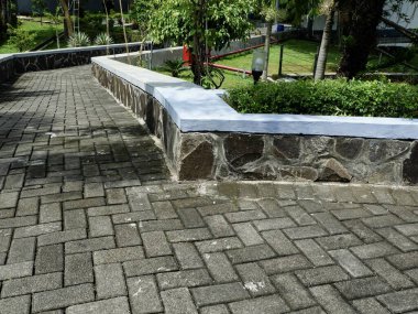 Modern garden walkway with grey herringbone paving blocks and a natural stone retaining wall. Landscaped outdoor path surrounded by lush green plants and trees in bright daylight.