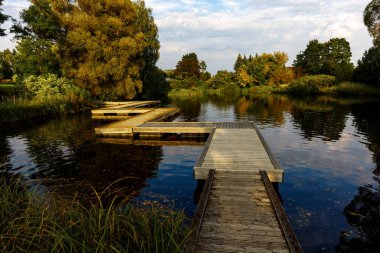 Gün batımında yemyeşil sonbahar ağaçlarıyla çevrili huzurlu bir gölet üzerinde tahta bir rıhtımın manzarası..