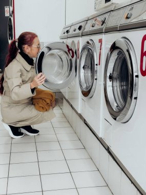 Uzun paltolu kırmızı at kuyruklu bir kadının modern çamaşırhanedeki endüstriyel çamaşır makinesini kontrol edişinin yan görüntüsü. Huzurlu şehir manzarası. Numarası: Beyaz iç mekan, temiz simetri, günlük görsel düzen.