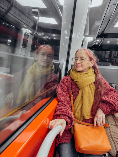 Traveler in Modern Train with Reflections A woman with pink hair and glasses sits on a train, wearing a mustard scarf and red sweater, holding an orange bag. Her reflection is visible in the window.