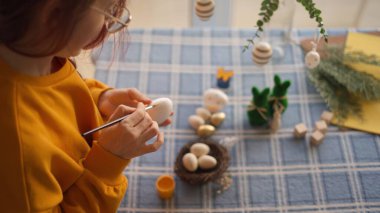 Masaya mavi kareli masa örtüsüyle Paskalya yumurtası çizen biri. Masa, yumurtalı bir yuva, küçük bir bitki ve tahta bloklar da dahil olmak üzere Paskalya temalı eşyalarla süslenmiş..