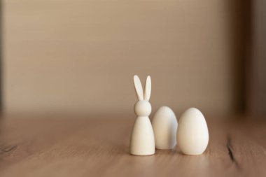 Minimum beyaz ahşap tavşancık figürü ve yumuşak bokeh arkaplanlı iki yumurta, tarafsız tonlar, kopyalama alanı.