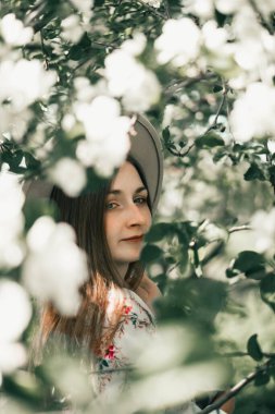 Yumuşak beyaz çiçekler ve canlı yeşil yapraklarla çevrili şık şapkalı bir kadının huzurlu portresi. Güneş ışığı onun sakin ifadesini vurguluyor, barışçıl ve zarif bir bahar atmosferi yaratıyor.