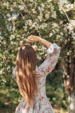 Dalgalı saçları olan zarif bir kadın, sakin bir bahar ortamında açan dallara uzanır. Yumuşak ışık çiçekli elbisesini ve doğayla olan barışçıl bağlantısını vurguluyor. Çiçekli Kadın