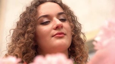 Kıvırcık saçlı, gülümseyen bir kadının çiçek açan güzelliğinin tadını çıkardığı yakın plan. Etrafı yumuşak pembe yapraklarla ve sıcak, doğal bir atmosferle çevrili, neşe ve takdir saçıyor. Yüksek kalite 4k