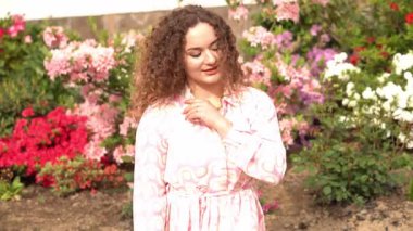 Kıvırcık saçlı neşeli bir kadın ve saçında pembe bir çiçek açan çiçeklerle güneş ışığının tadını çıkarıyor. Onun gevşek ifadesi ve yumuşak gülümsemesi sıcaklık ve mutluluk saçıyor. Yüksek kalite 4k