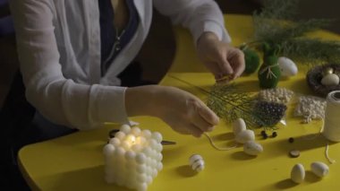 Sarı masada paskalya yumurtaları, yeşillik, sicim, süslemeler ve parıldayan bir küp lamba yapan eller. Sarı masada Paskalya yumurtası süslemeleri, sarı masada Paskalya yumurtası süslemeleri.